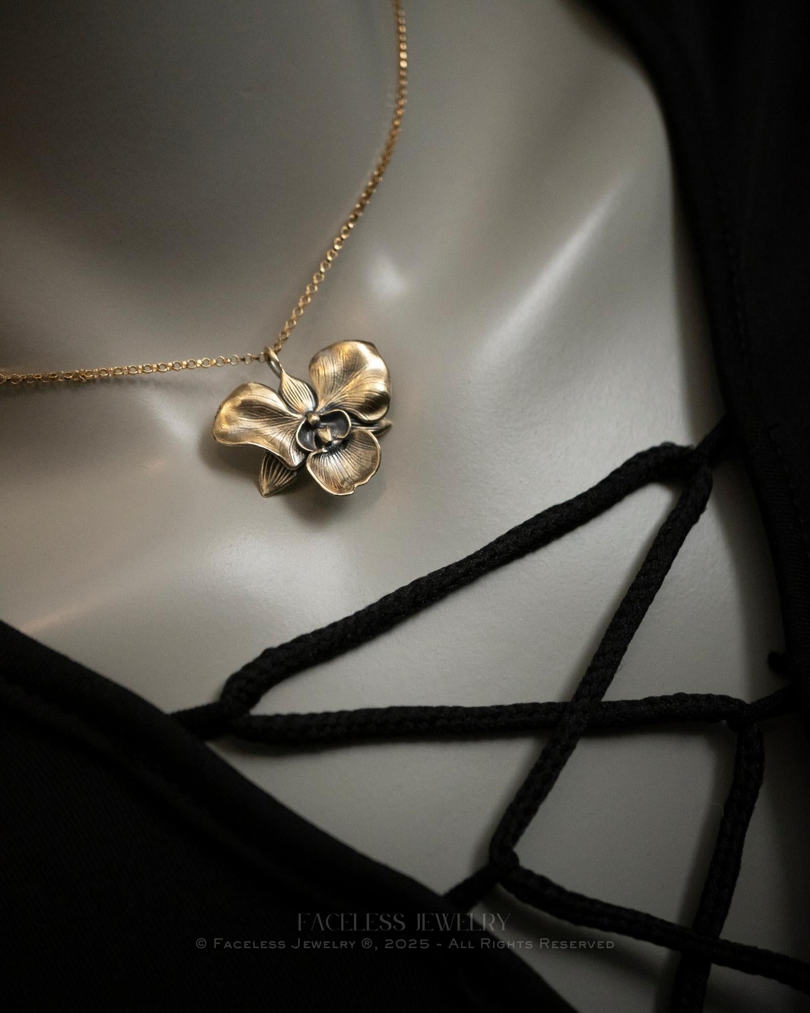The Golden Orchid pendant by Faceless Jewelry displayed on a mannequin wearing a black lace-up top, featuring a 14Kt gold orchid flower pendant with soft, detailed petals and a brushed finish on a fine gold chain resting near the collarbone.
