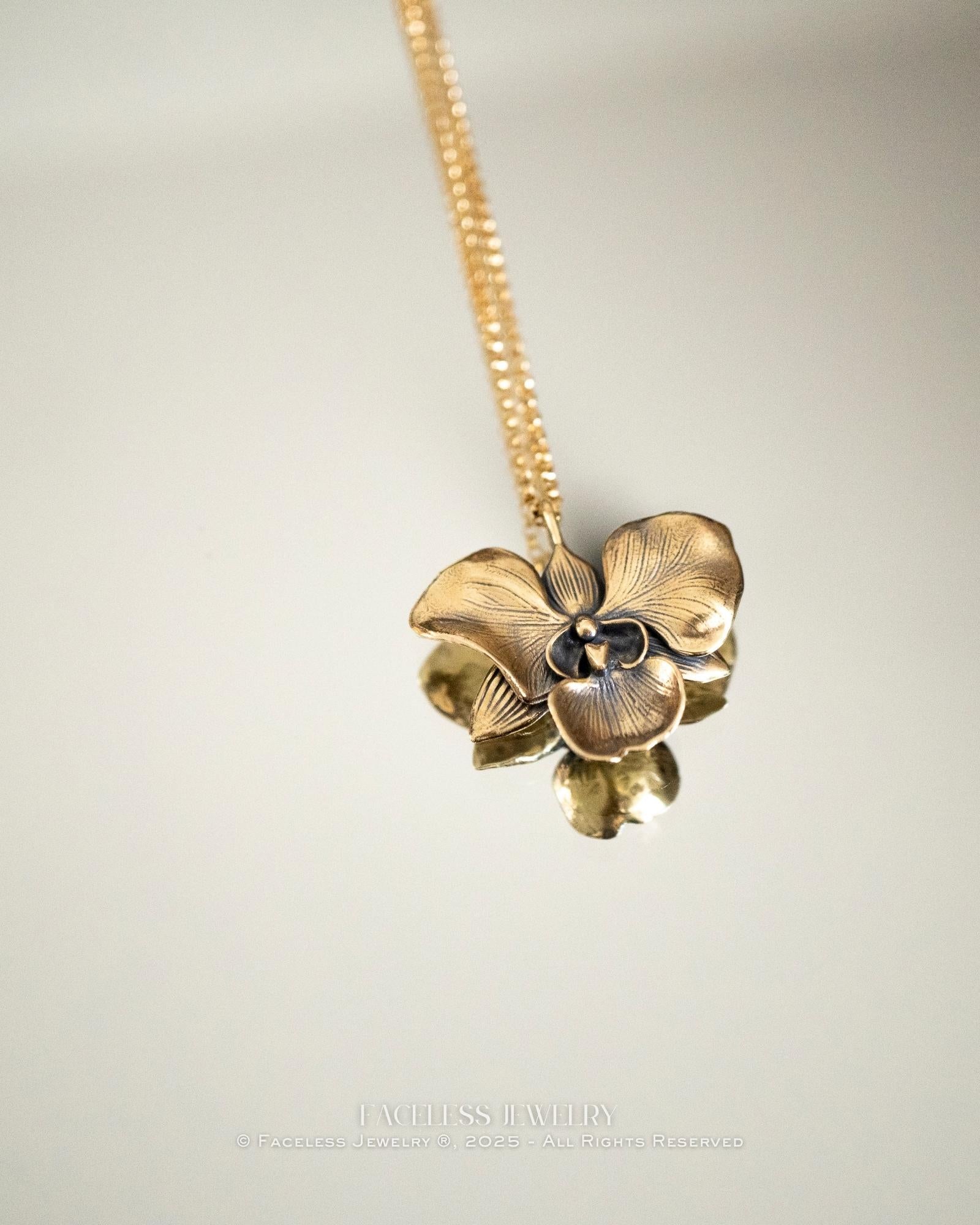 The Golden Orchid pendant by Faceless Jewelry resting on a mirror, featuring a 14Kt gold orchid flower with detailed petals and a brushed finish, its reflection softly visible beneath the gold chain.