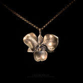A close-up of the Golden Orchid pendant by Faceless Jewelry, a detailed 14Kt gold orchid flower with softly curved petals and fine vein textures, suspended on a gold rolo chain against a black background.