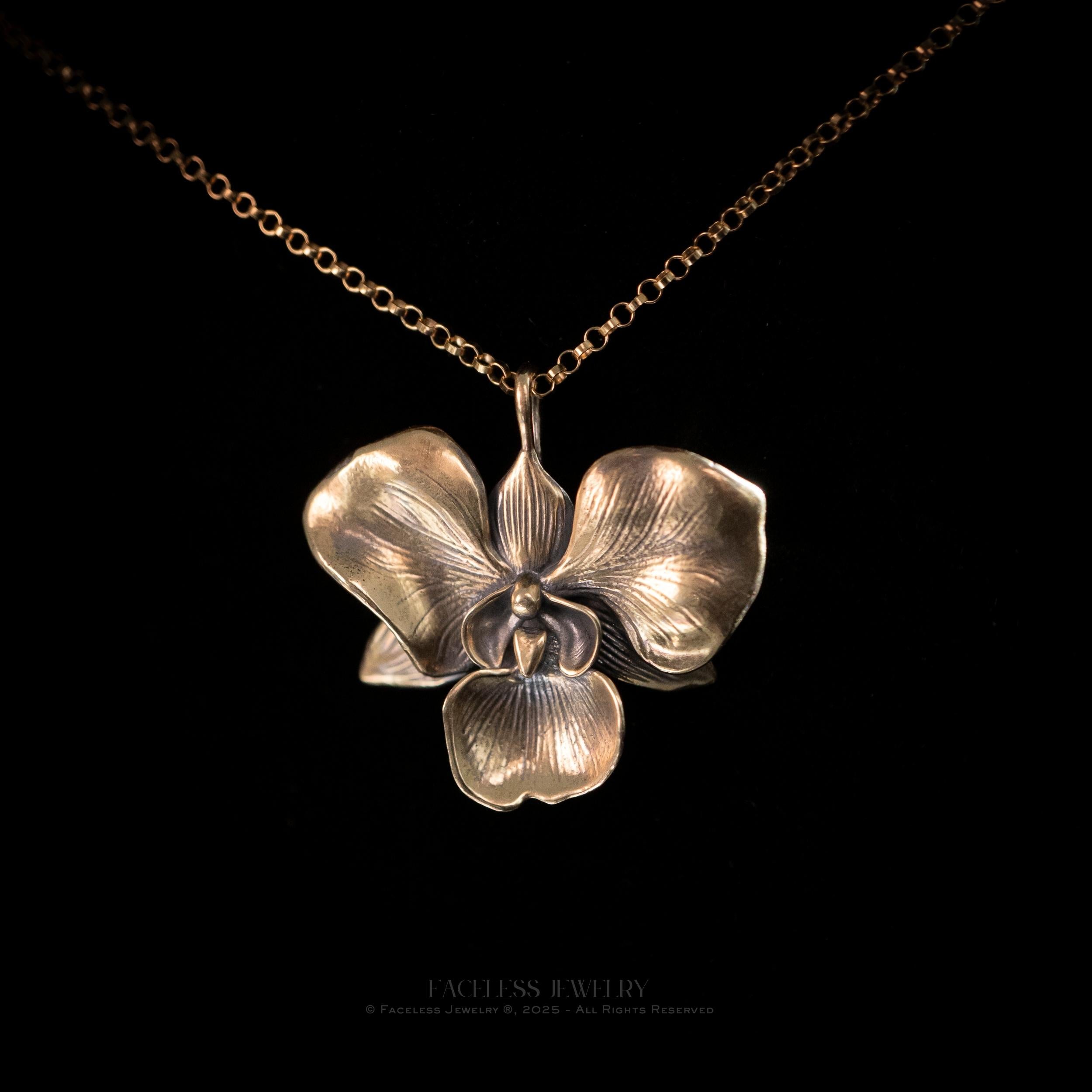 A close-up of the Golden Orchid pendant by Faceless Jewelry, a detailed 14Kt gold orchid flower with softly curved petals and fine vein textures, suspended on a gold rolo chain against a black background.