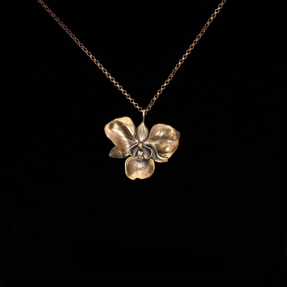 The Golden Orchid pendant by Faceless Jewelry displayed against a black background, featuring a 14Kt gold orchid flower with finely detailed petals and a warm, polished finish, hanging from a delicate gold rolo chain.