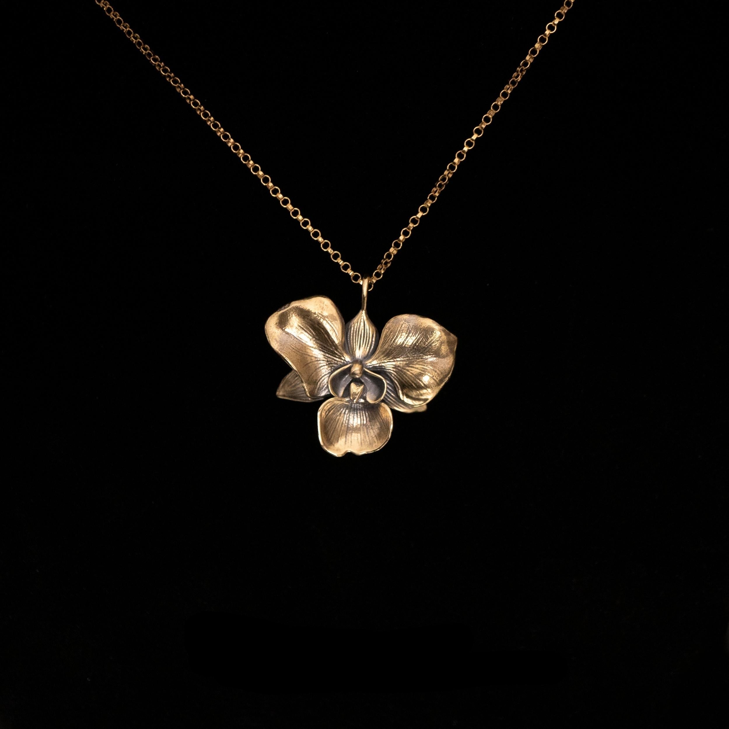 The Golden Orchid pendant by Faceless Jewelry displayed against a black background, featuring a 14Kt gold orchid flower with finely detailed petals and a warm, polished finish, hanging from a delicate gold rolo chain.