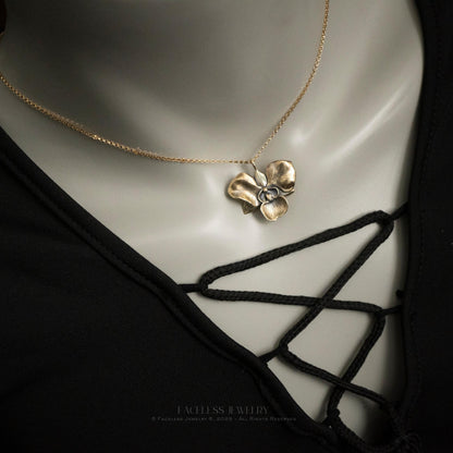 The Golden Orchid pendant by Faceless Jewelry shown on a mannequin wearing a black lace-up top, featuring a 14Kt gold orchid flower with detailed petals suspended from a fine gold chain resting near the collarbone.