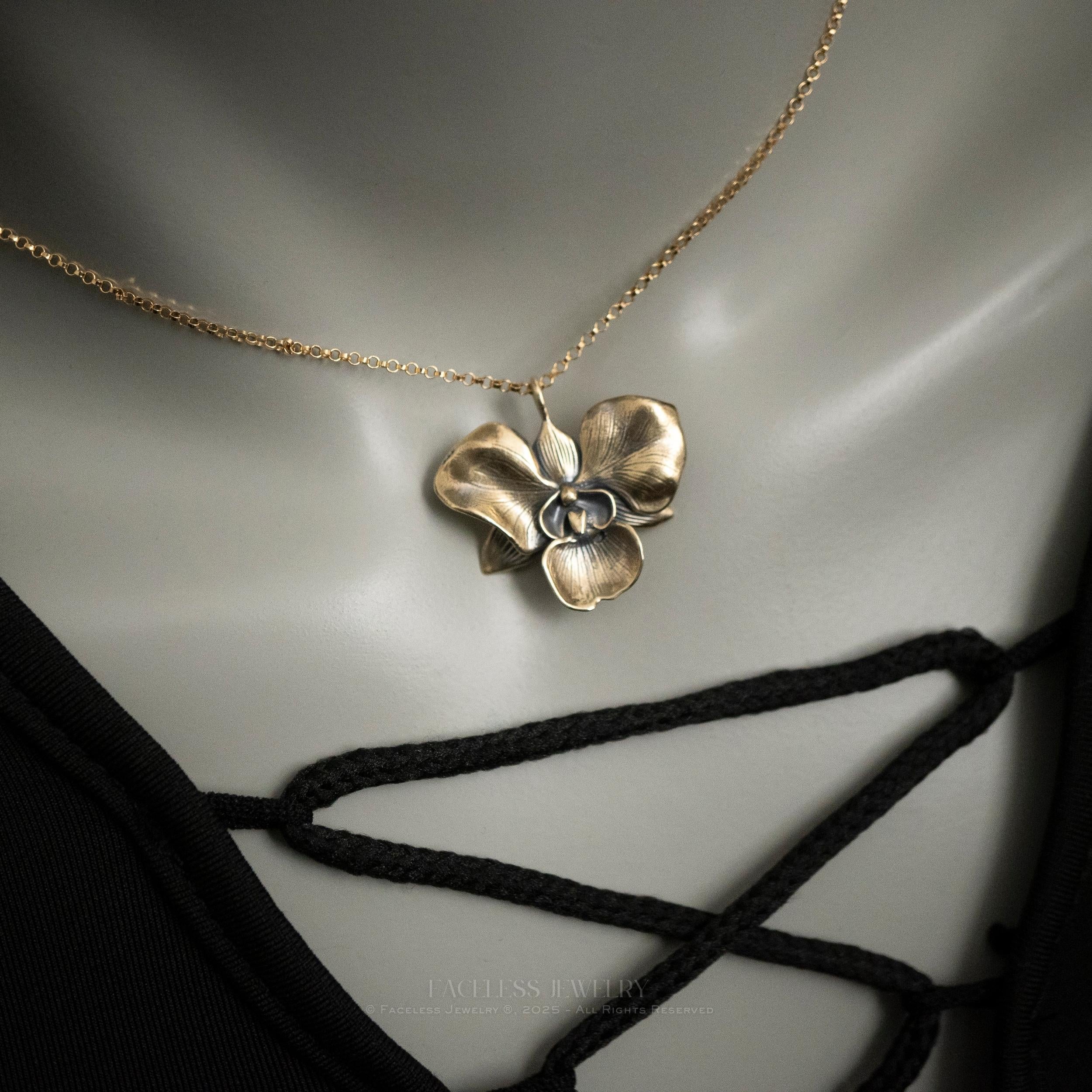 The Golden Orchid pendant by Faceless Jewelry displayed on a mannequin wearing a black lace-up top, featuring a 14Kt gold orchid flower pendant with soft, detailed petals and a brushed finish on a fine gold chain resting near the collarbone.
