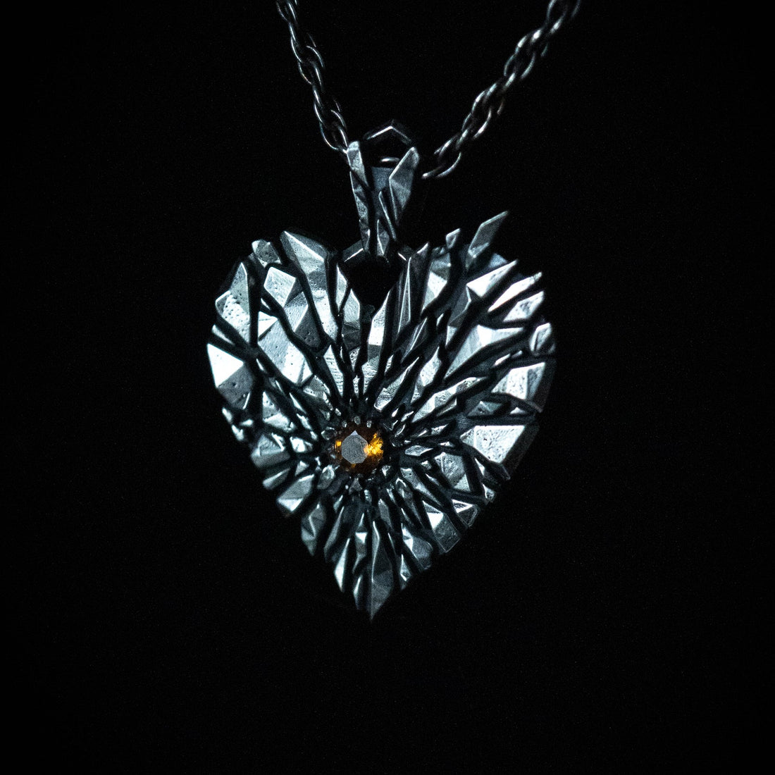 A heart-shaped silver pendant necklace with a faceted, fractured design and an orange gemstone at the center, suspended from a dark rope chain against a solid black background.