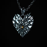 A heart-shaped silver pendant necklace with a faceted, fractured design and an orange gemstone at the center, suspended from a dark rope chain against a solid black background.