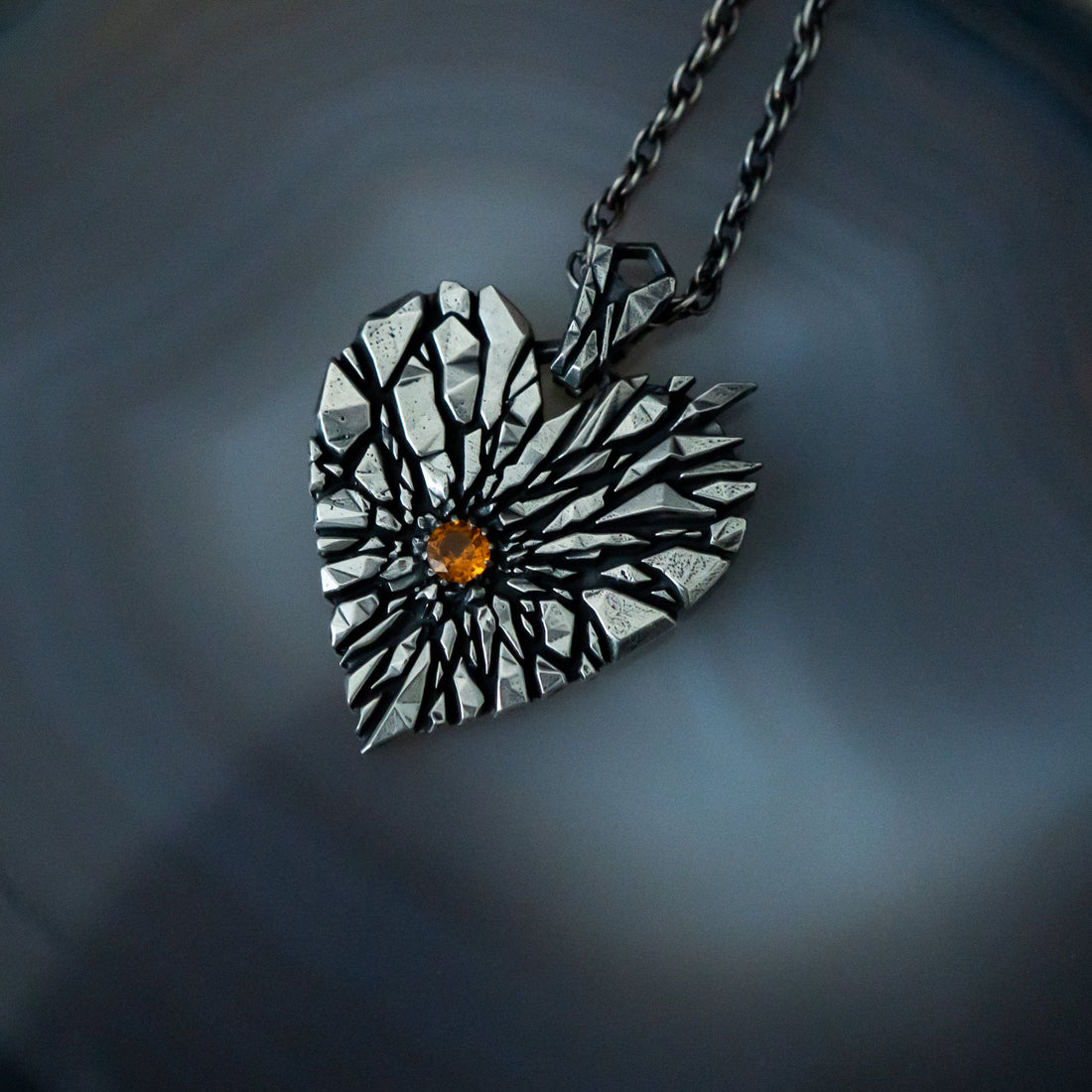 A heart-shaped pendant necklace with a textured silver and black sunburst design and a small amber gemstone in the center, resting on a dark agate surface.