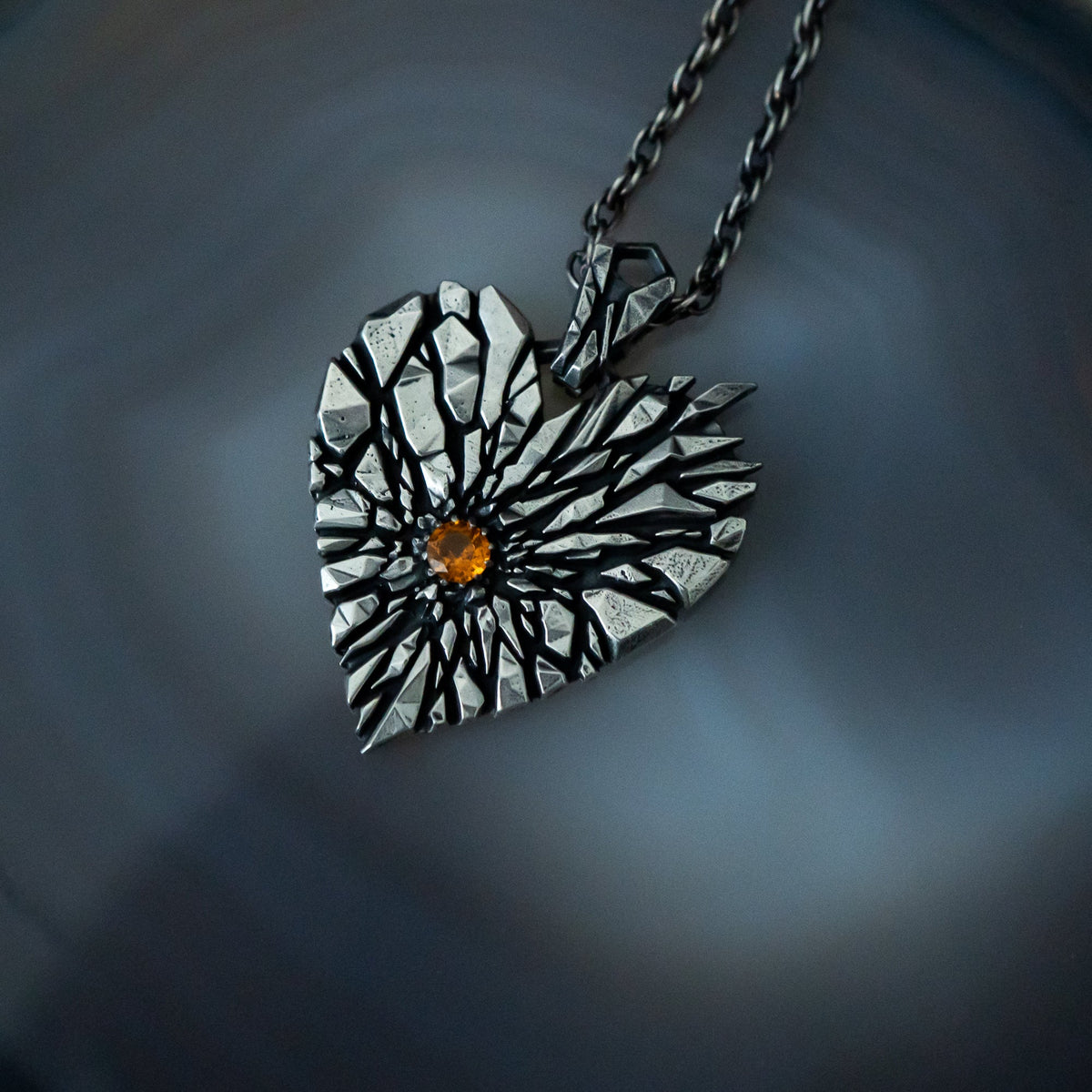 A heart-shaped pendant necklace with a textured silver and black sunburst design and a small amber gemstone in the center, resting on a dark agate surface.