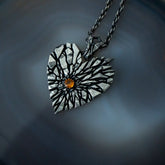 A heart-shaped pendant necklace with a textured silver and black sunburst design and a small amber gemstone in the center, resting on a dark agate surface.