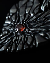 A close-up of a black faceted ring with a central round orange gemstone, photographed against a black background.