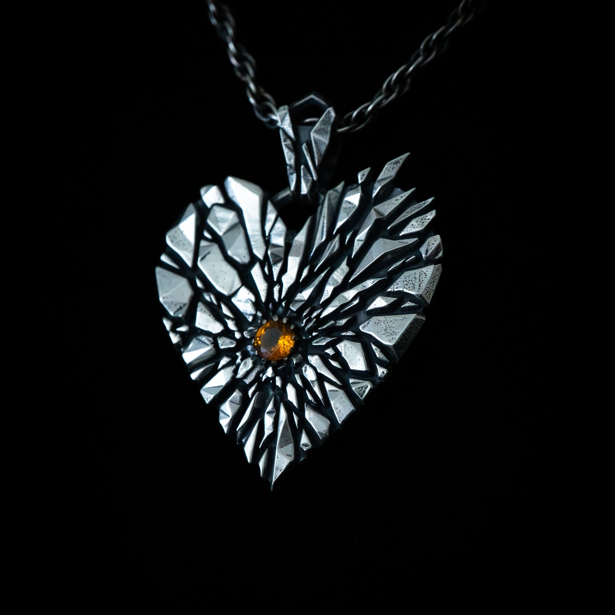 A heart-shaped pendant necklace with a textured, cracked metallic finish and a small amber gemstone at the center, suspended on a dark chain against a solid black background.