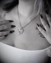 A close-up lifestyle shot of a model wearing a silver heart-shaped pendant necklace with a small orange gemstone accent. The image is in black and white, highlighting the model&