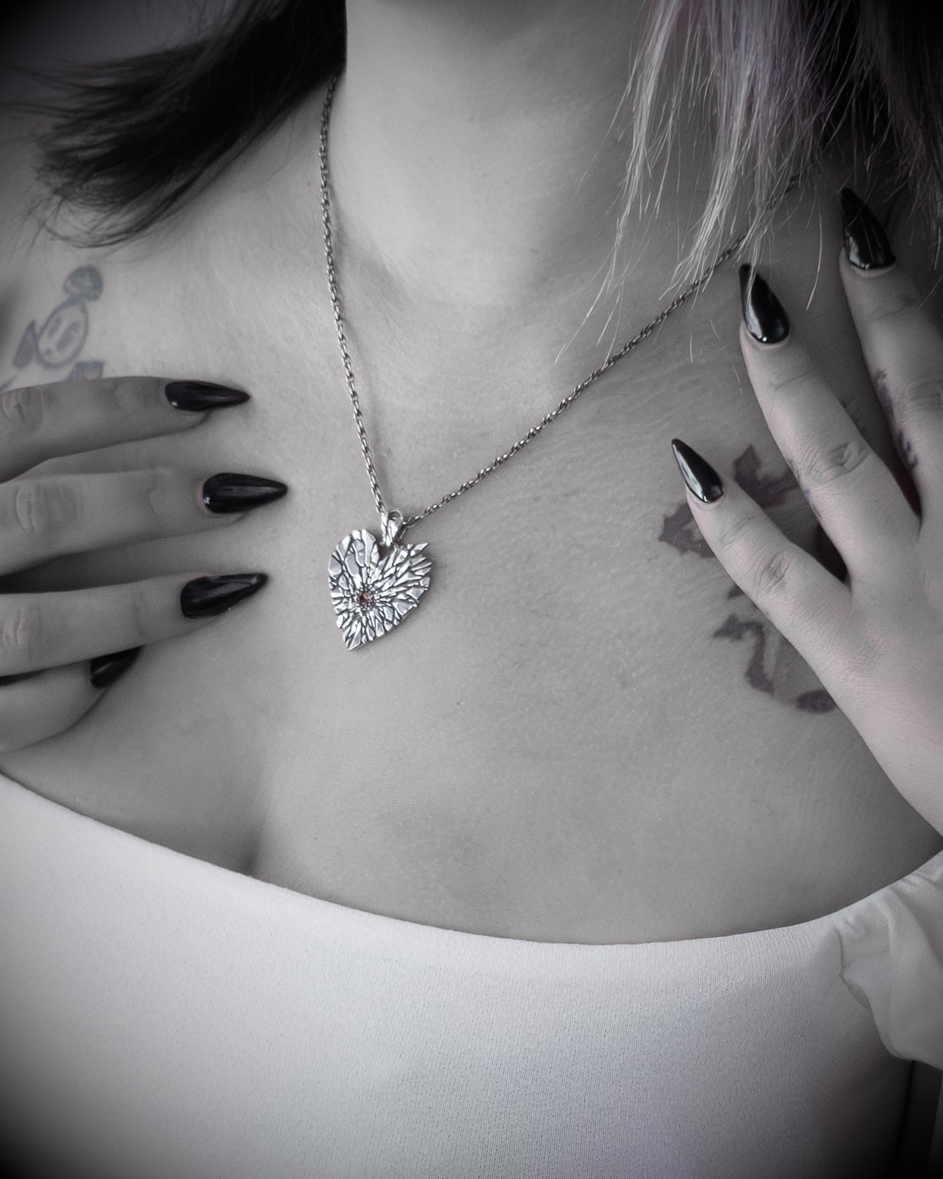 A close-up lifestyle shot of a model wearing a silver heart-shaped pendant necklace with a small orange gemstone accent. The image is in black and white, highlighting the model&