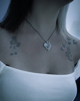 A close-up lifestyle shot of a person wearing a silver heart-shaped pendant necklace on a twisted chain, styled with a white top and visible tattoos on the shoulders.
