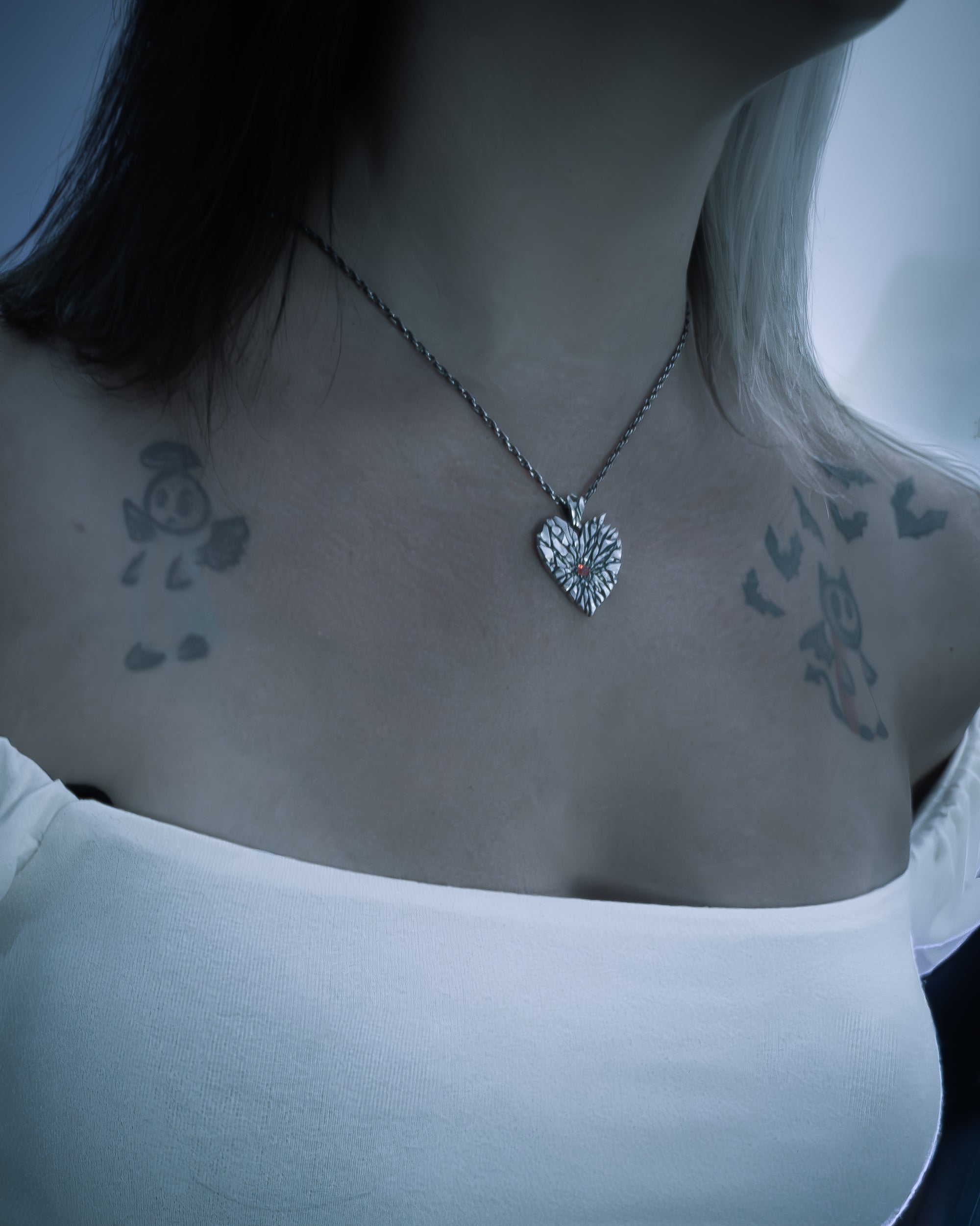 A close-up lifestyle shot of a person wearing a silver heart-shaped pendant necklace on a twisted chain, styled with a white top and visible tattoos on the shoulders.