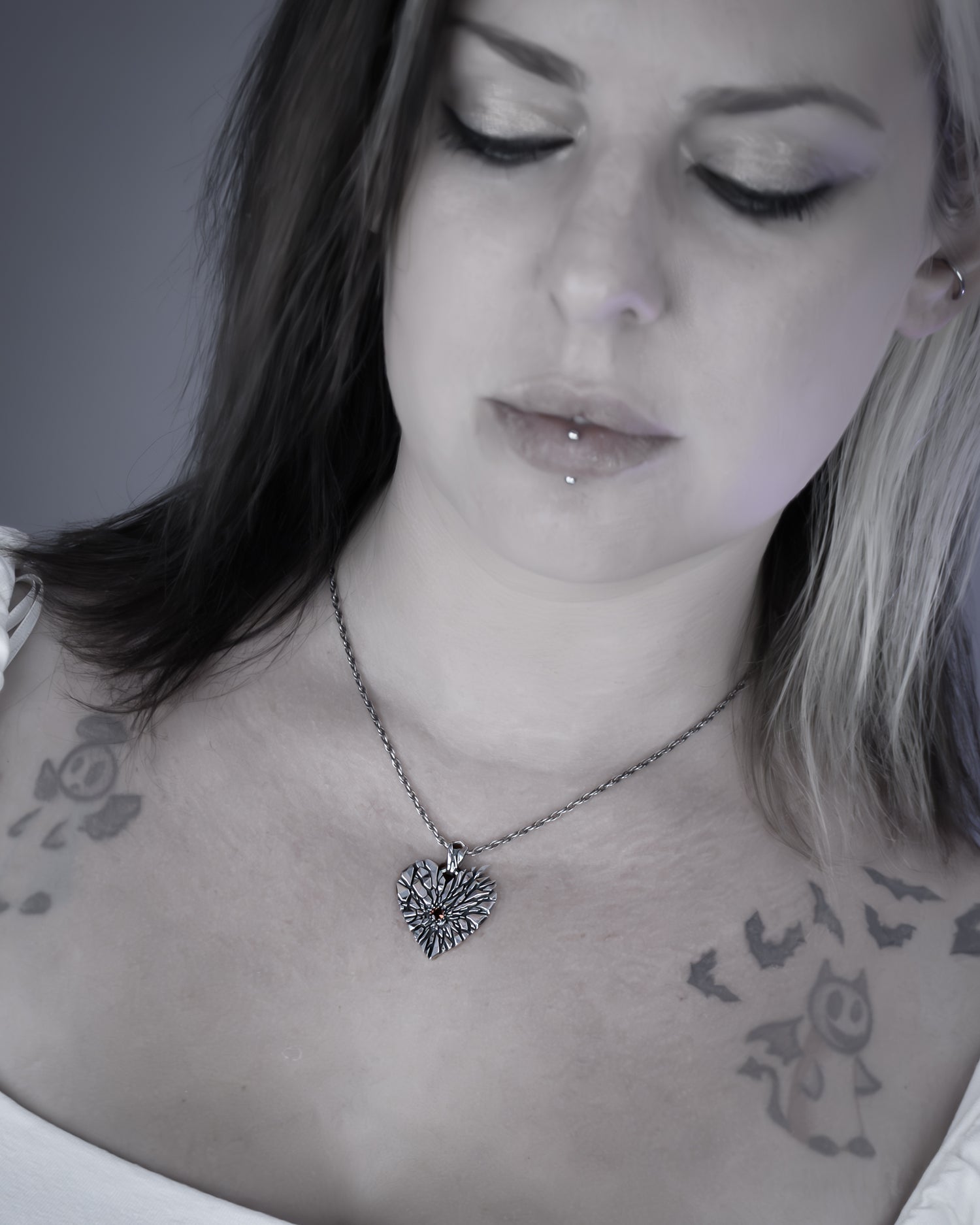 A close-up lifestyle shot of a woman wearing a silver heart-shaped pendant necklace with a fractured design, set against a soft gray background.