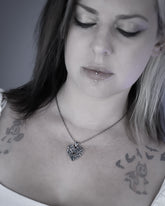A close-up lifestyle shot of a woman wearing a silver heart-shaped pendant necklace with a fractured design, set against a soft gray background.